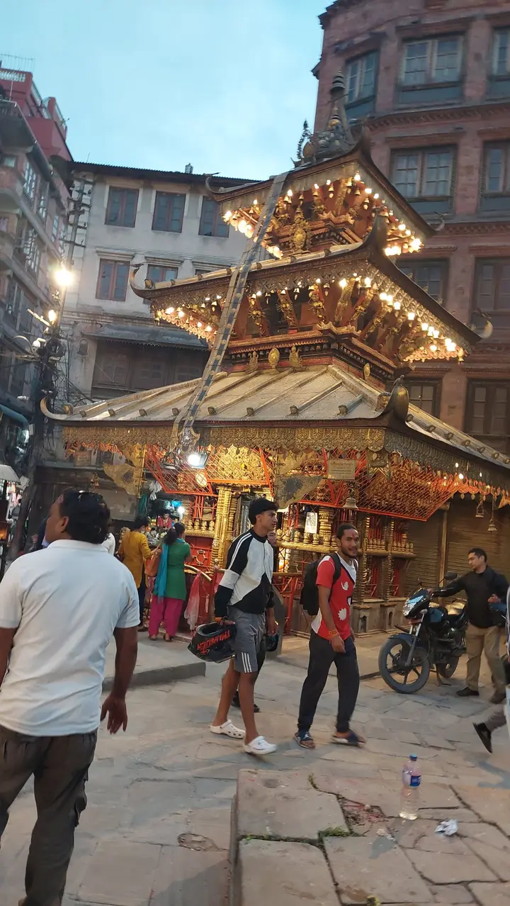 A gilded three-tiered pagoda temple lit with bulbs at dusk, with people walking past in the old town