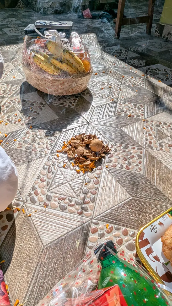 Cracked walnut shells and marigold petals on a tiled floor, with a wrapped fruit basket and gifts nearby and incense smoke in the air