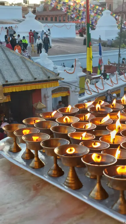 Rows of lit brass butter lamps on a tray, with the white stupa terrace, prayer flags and visitors behind