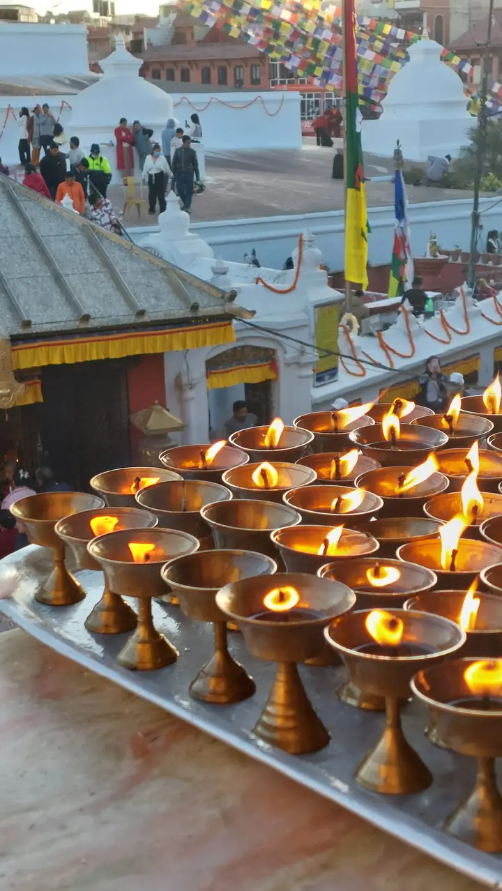Rows of lit brass butter lamps on a tray, with the white stupa terrace, prayer flags and visitors behind