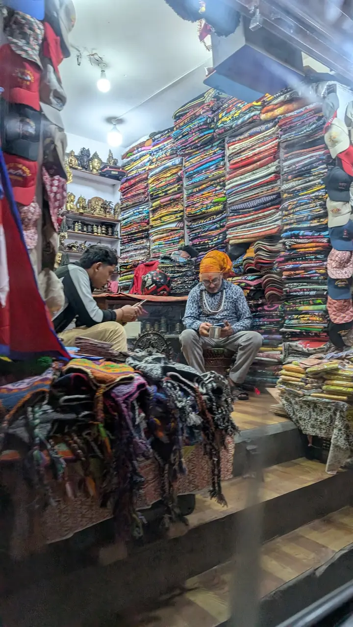 A small shop stacked to the ceiling with colourful folded cloth, brass statues on the shelves; the shopkeeper wears a tika and an orange head cloth