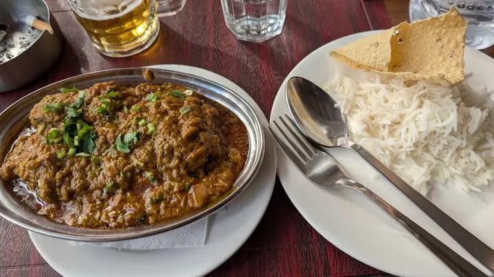 A steel dish of curry garnished with coriander, next to a plate of rice with papad, and a glass of beer