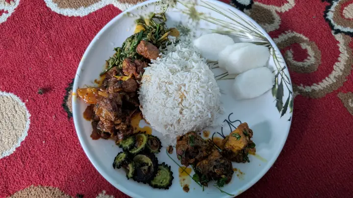 A plate of rice with meat curry, fried greens, bitter gourd, spiced meat and slices of radish, on a red rug