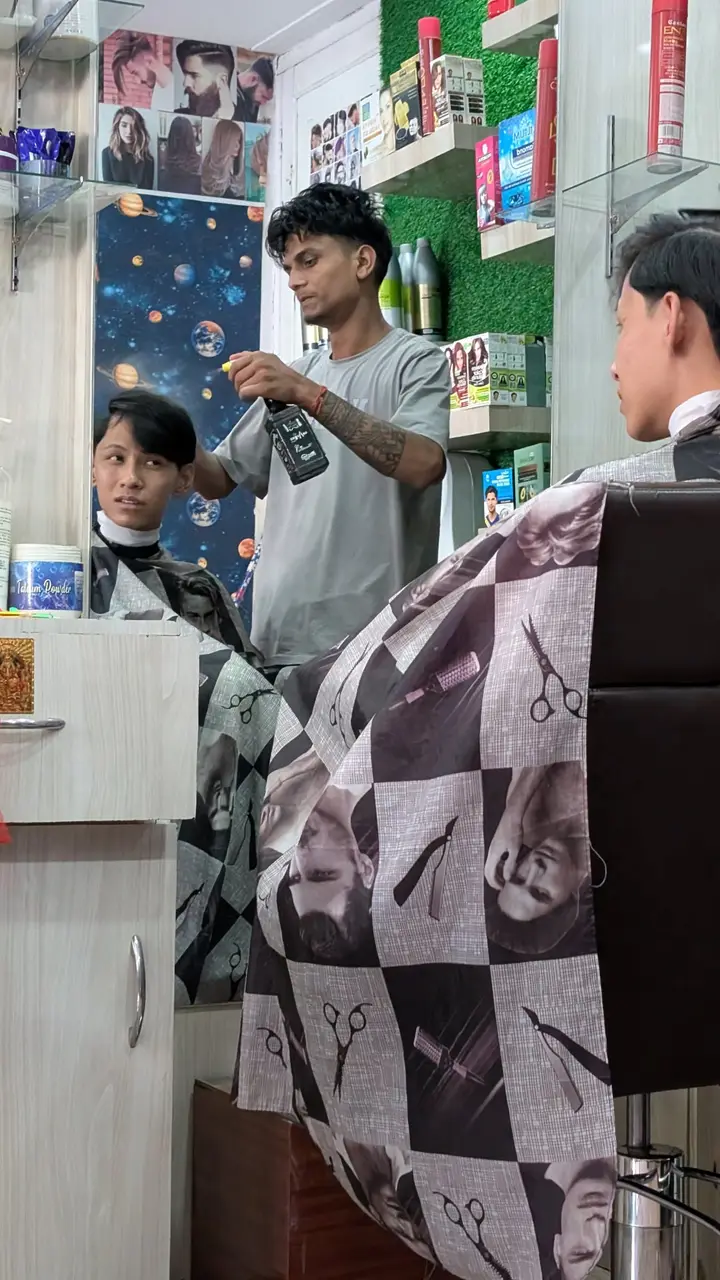 A barber sprays water on a young man's hair in a small salon, with another customer in the foreground chair