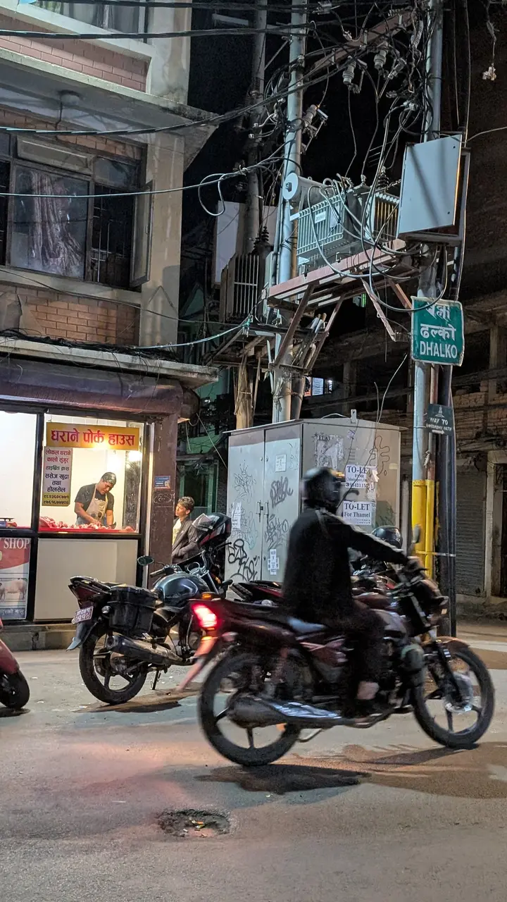 A butcher shop lit up at night under a tangle of power lines, a motorbike passing and a street sign reading Dhalko