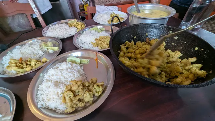 Steel plates of rice with cauliflower and potato curry, a wok of curry and a pot of dal on a table