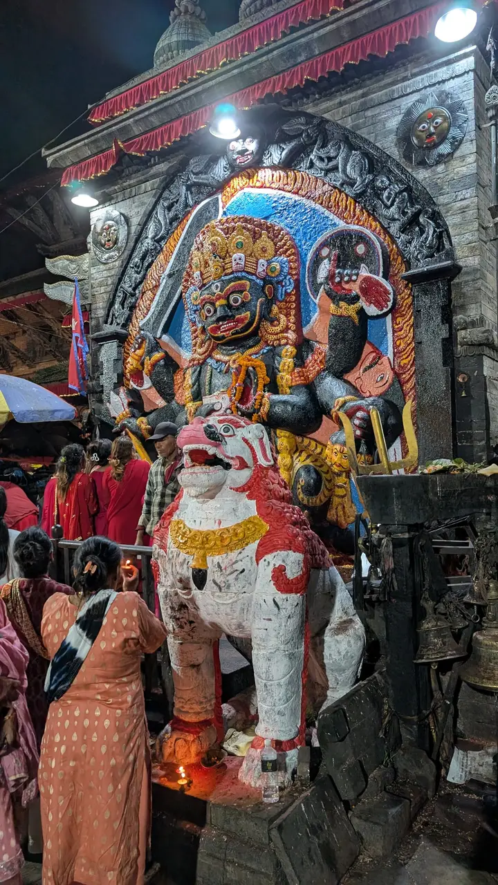 Women holding small oil lamps worship in front of Kal Bhairav and a white and red stone lion