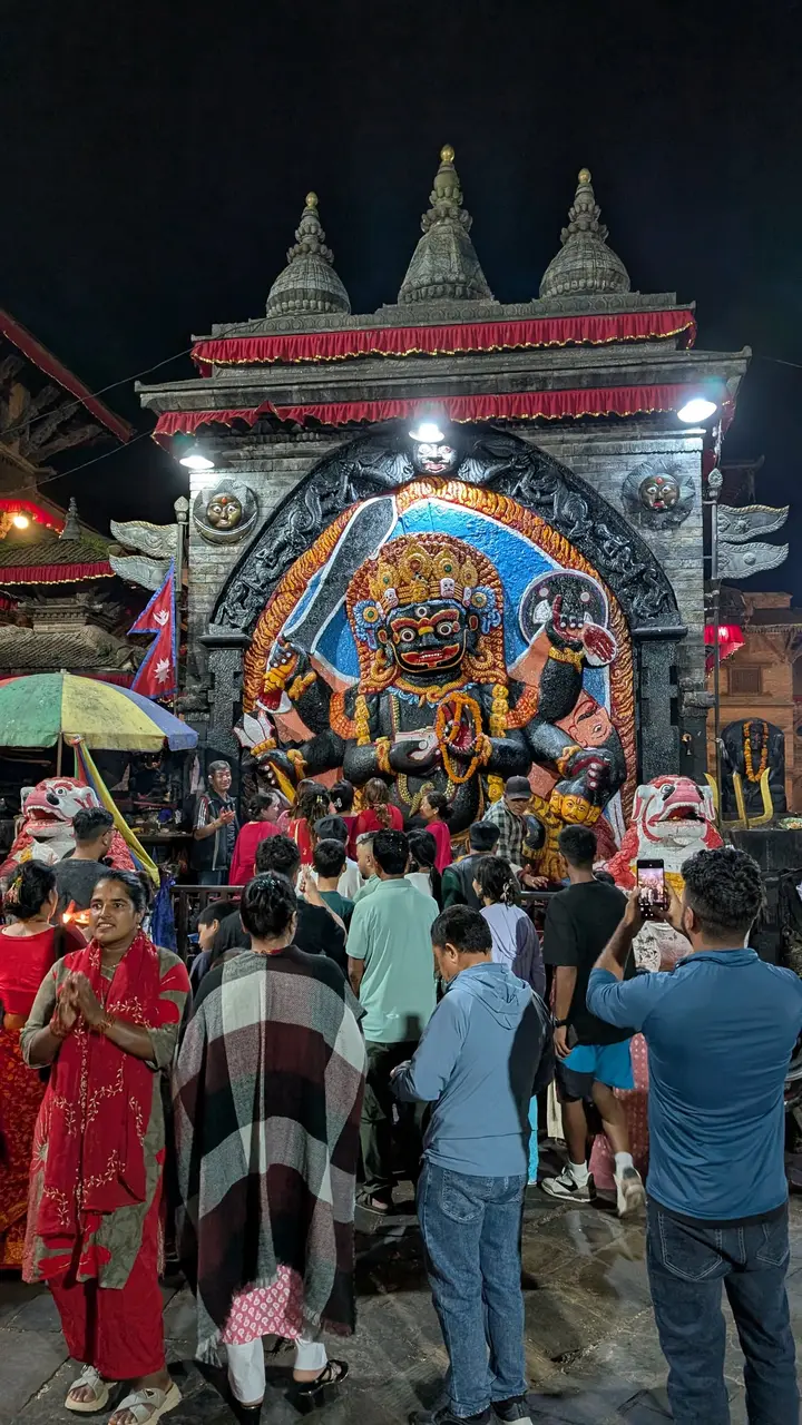 The large painted stone image of Kal Bhairav, black with a gold crown, lit at night, with a crowd of worshippers in front