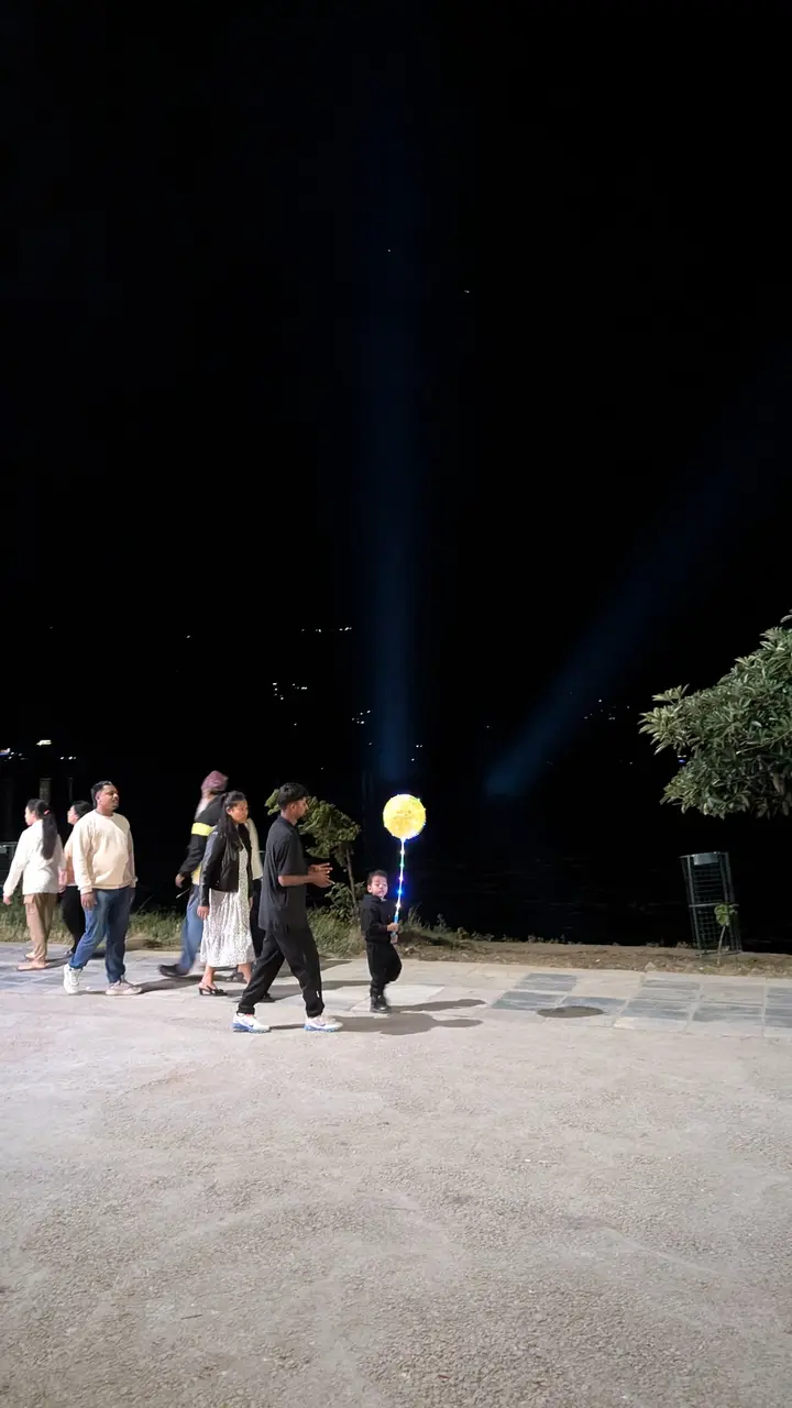 Families walk along a lakeside path at night; a small child carries a glowing balloon on a stick