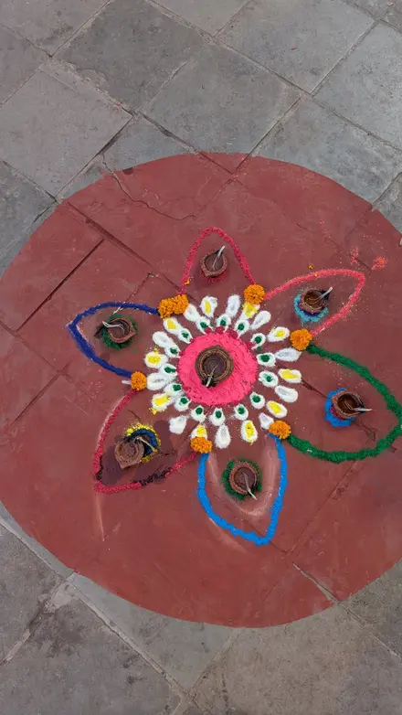 A flower-shaped rangoli of coloured powder on a red clay circle, with clay diyo lamps and marigolds on its petals