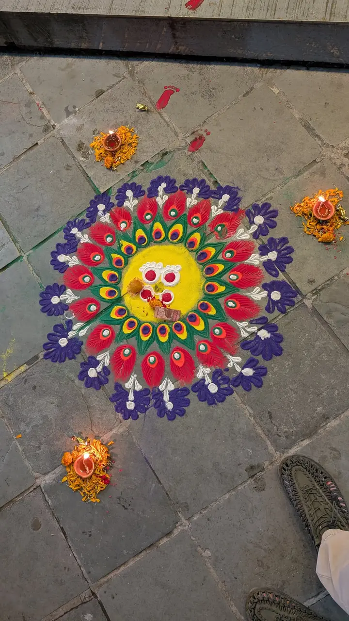 A bright rangoli with peacock-feather patterns and lit diyo lamps, with small red footprints painted beside it leading to the door