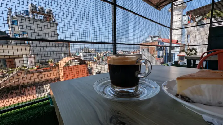 A glass cup of black coffee and a slice of cake on a rooftop café table, with Kathmandu rooftops and hills behind a mesh fence