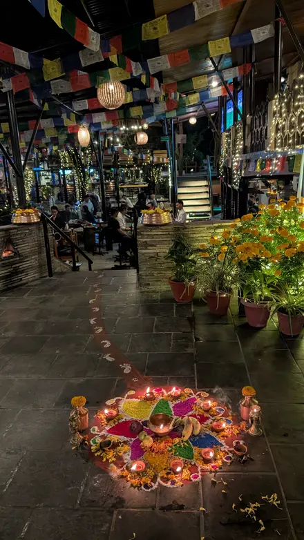 A rangoli with oil lamps, brass pots and fruit on a stone floor; painted white footprints lead from it into a courtyard hung with prayer flags and fairy lights