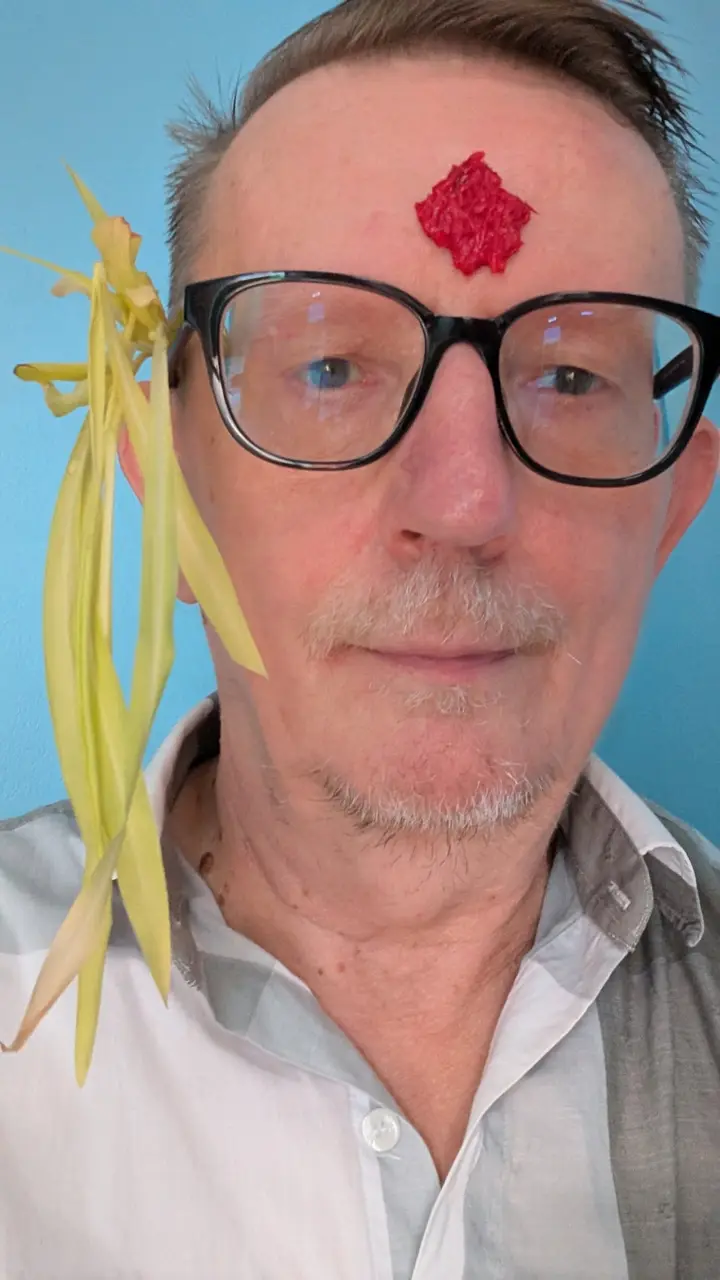 A man in glasses with a large red tika on his forehead and yellow jamara shoots tucked behind his ear