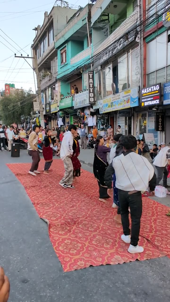 People in traditional dress dance on red carpets laid along a shopping street, watched by a crowd